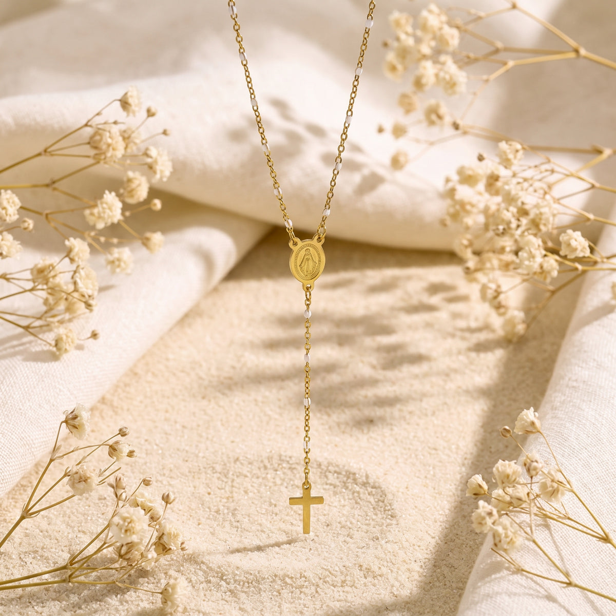 Gold-tone rosary lariat necklace displayed vertically over sand, white fabric, and dried flowers.