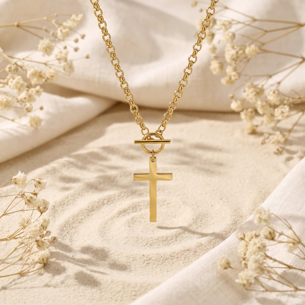 Gold-tone toggle cross necklace suspended vertically above rippled sand and dried flowers.