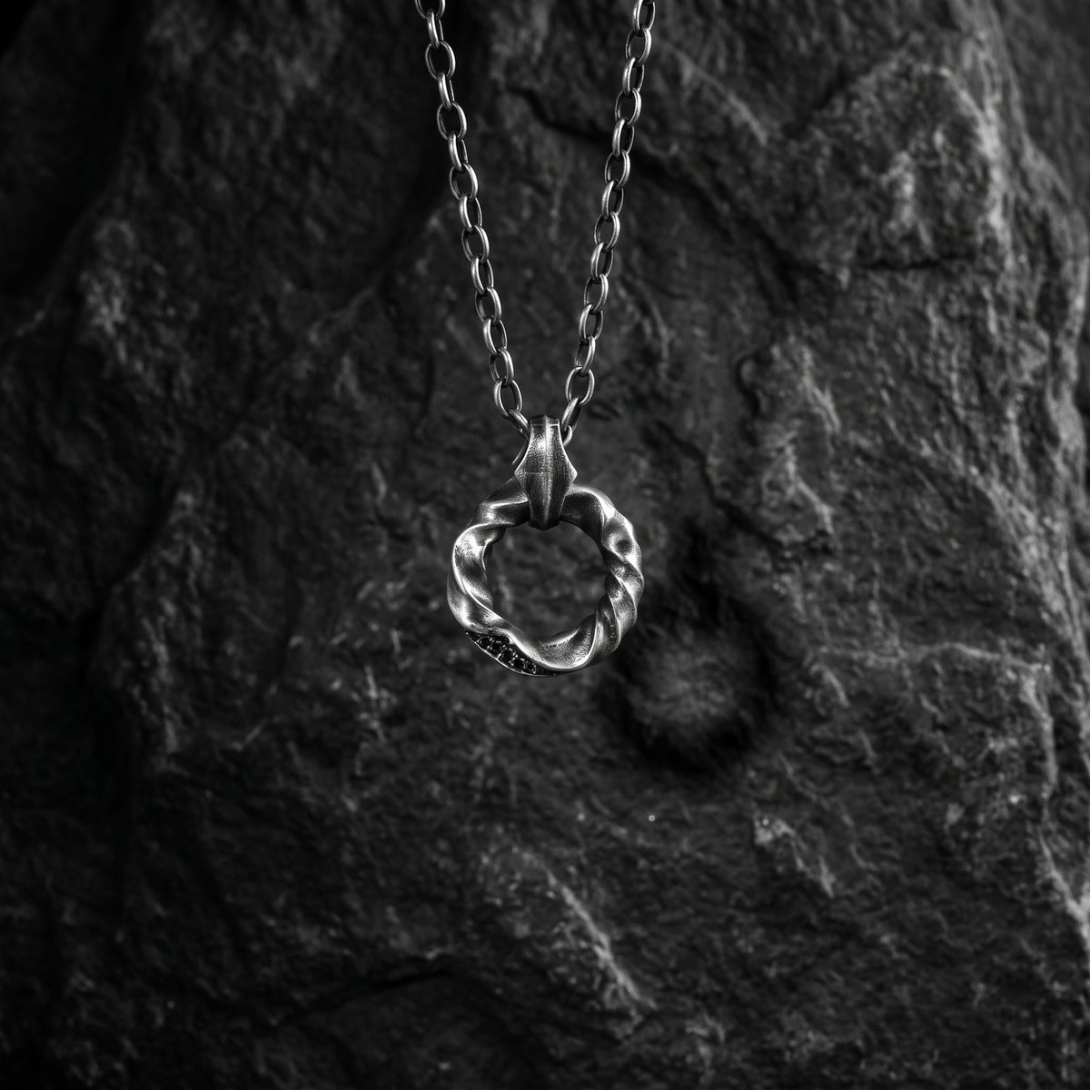 Silver twisted circle pendant hanging vertically from its chain against a dark textured rock backdrop.