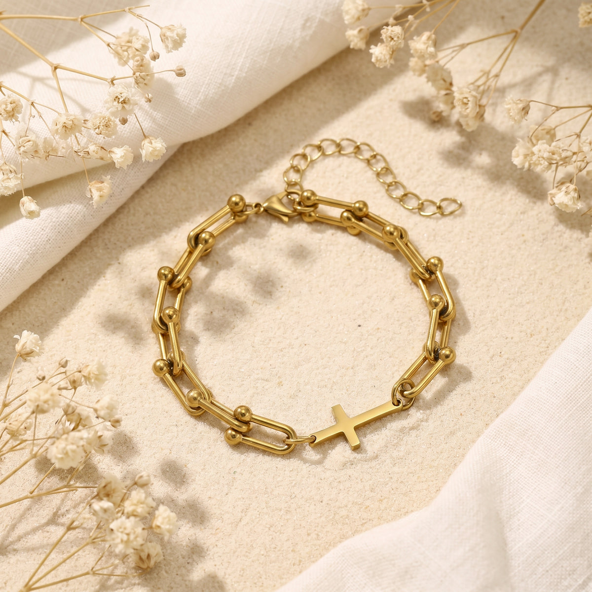 Alternate flat-lay view of the gold-tone cross bracelet on sand surrounded by white dried flowers and cloth.