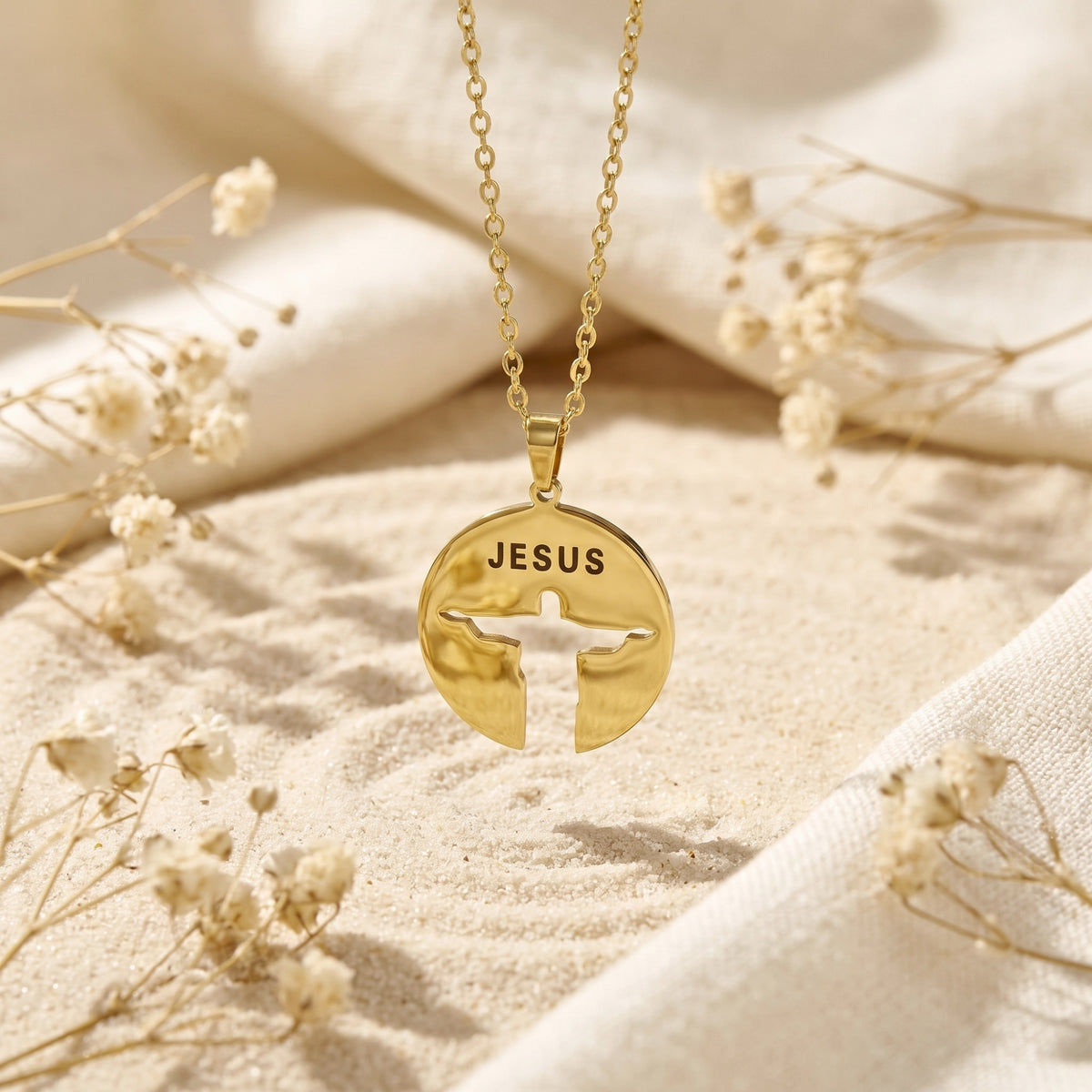 Gold-tone disc pendant with JESUS text and a Christ silhouette cutout, suspended over sand and dried flowers.