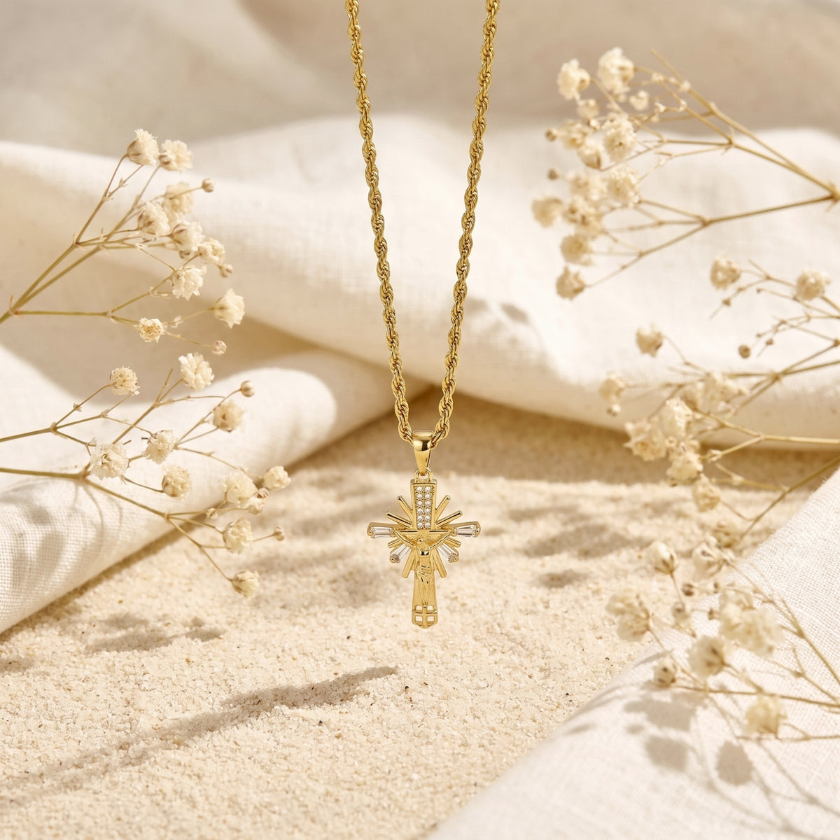 Gold-toned sunburst cross pendant suspended above sand, flanked by soft white linen and dried flowers.