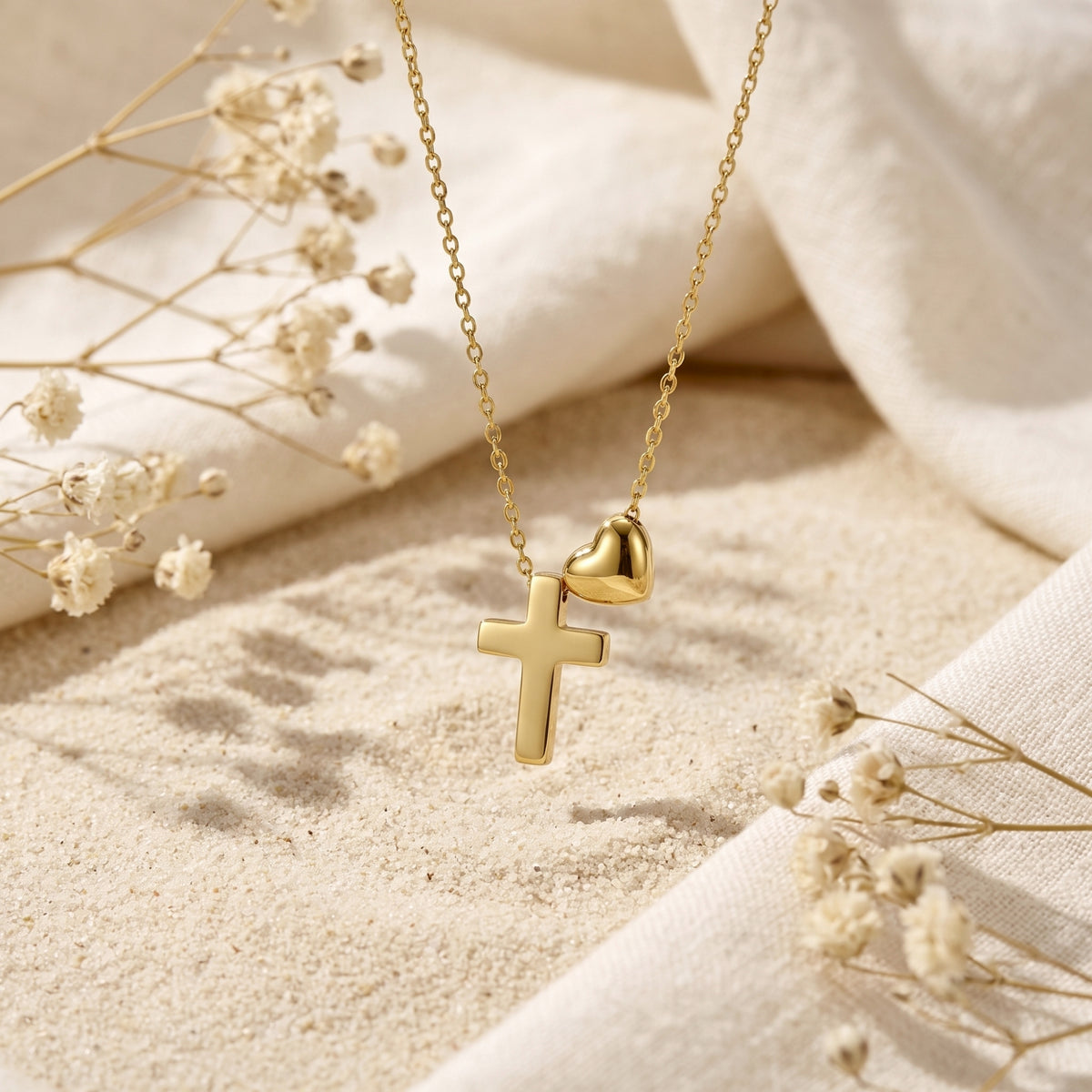 Gold-tone cross and heart necklace suspended above light sand with draped white fabric and dried flowers.