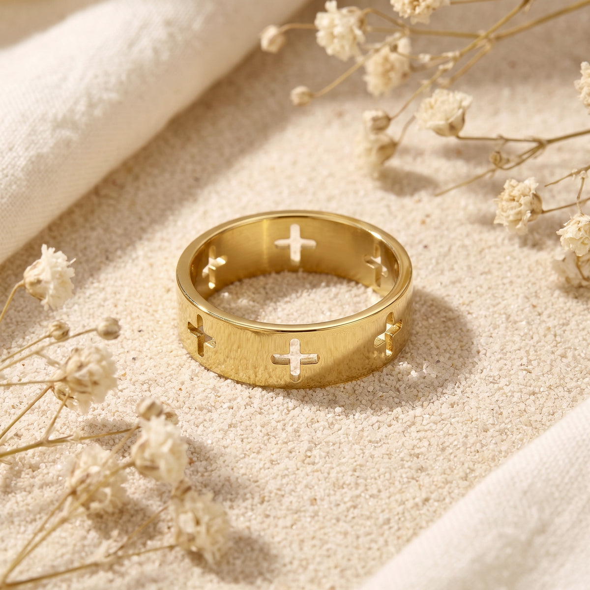 Gold-tone cross cutout band ring lying on sand, with its interior and front cross motifs visible.