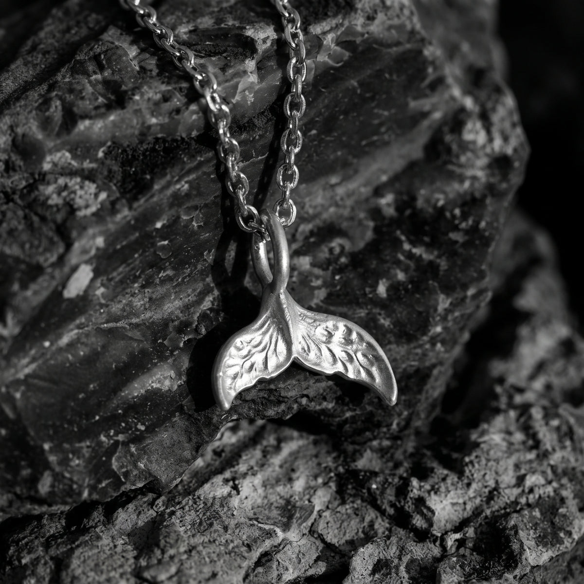 Front-facing close-up of a textured silver whale tail pendant and chain resting on rough dark rock.