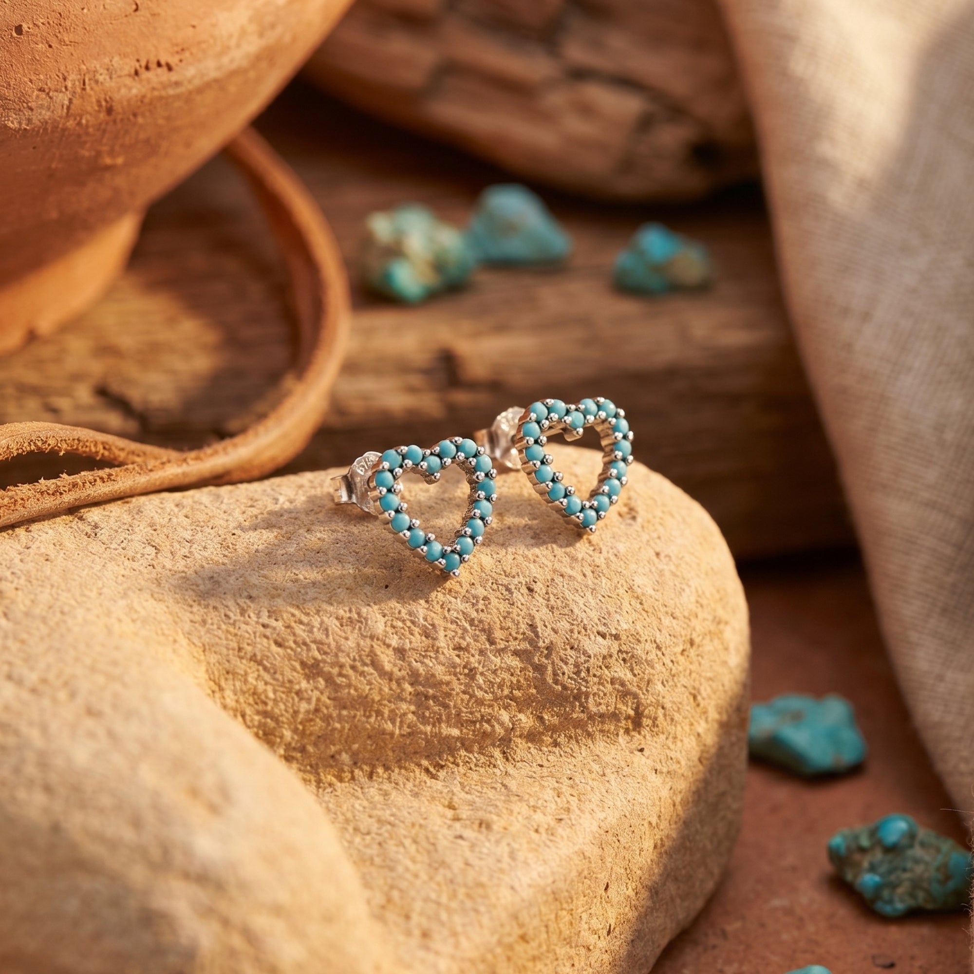 Pair of silver open-heart turquoise earrings displayed on a textured rock in a warm desert environment.