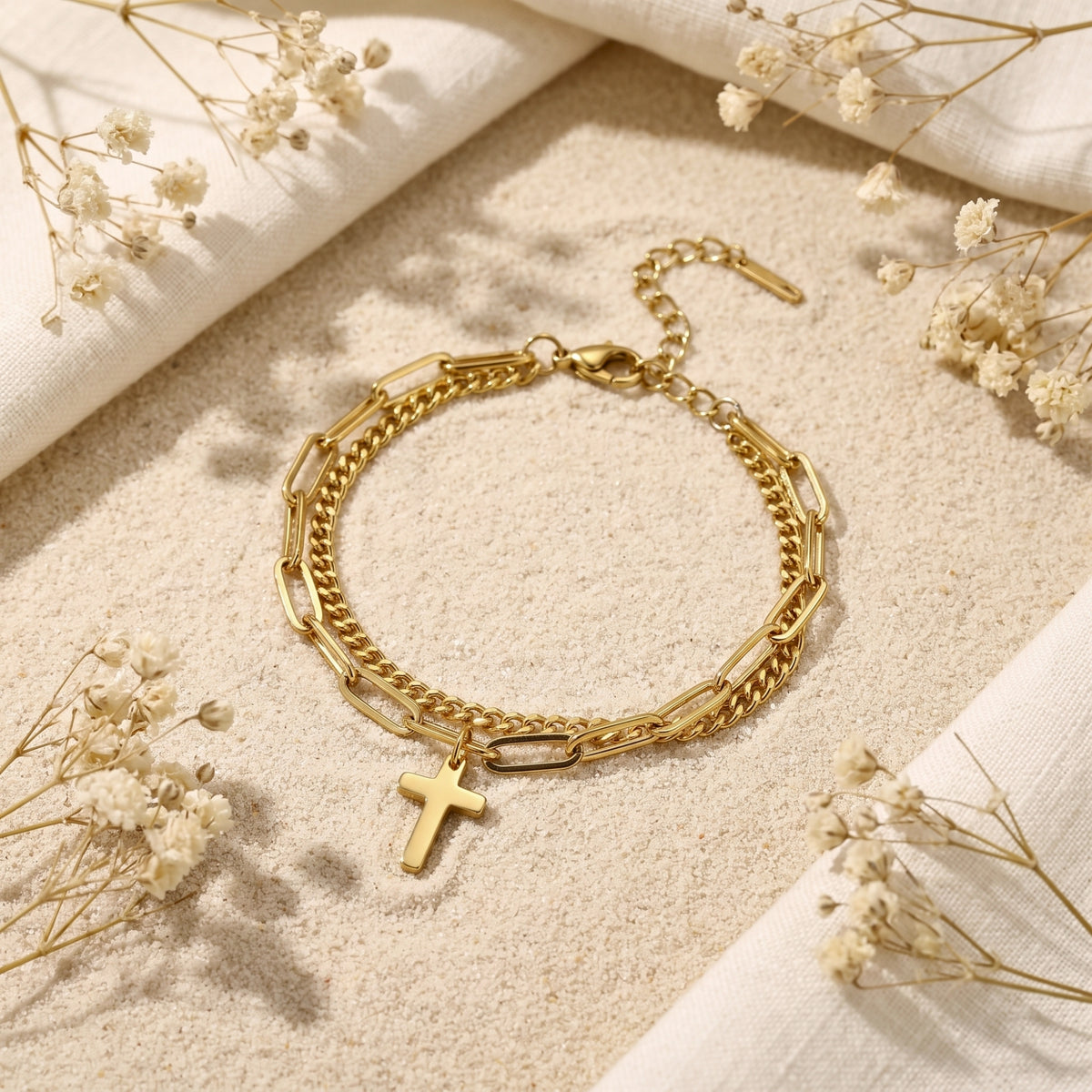 Gold-tone layered cross bracelet resting on fine sand, bordered by linen fabric and dried white flowers.