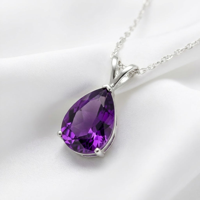 Close-up of a pear-cut purple moissanite necklace in a silver V-bale setting on white fabric.