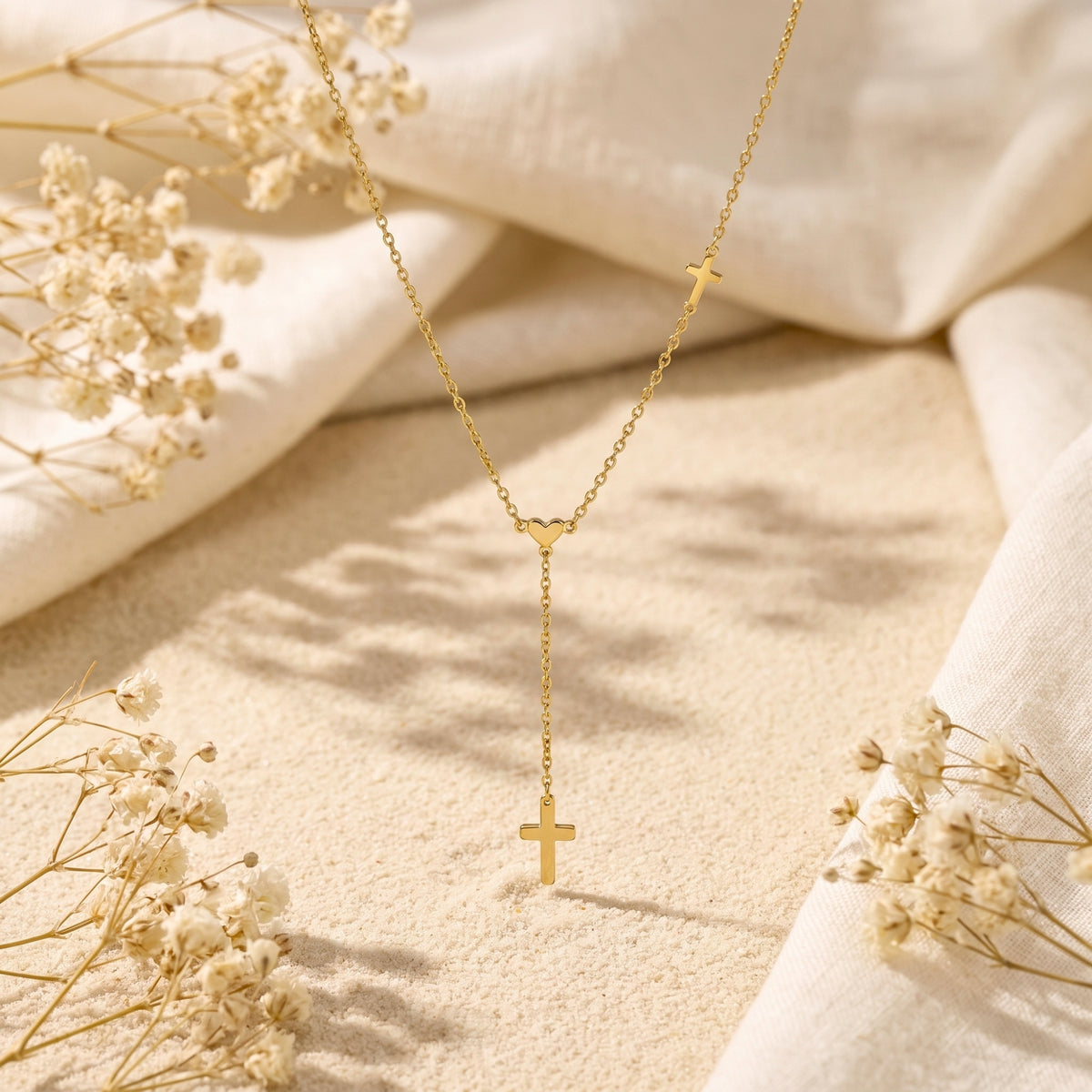 Vertical flat lay of the gold-tone lariat necklace on sand, bordered by white cloth and baby's-breath flowers.
