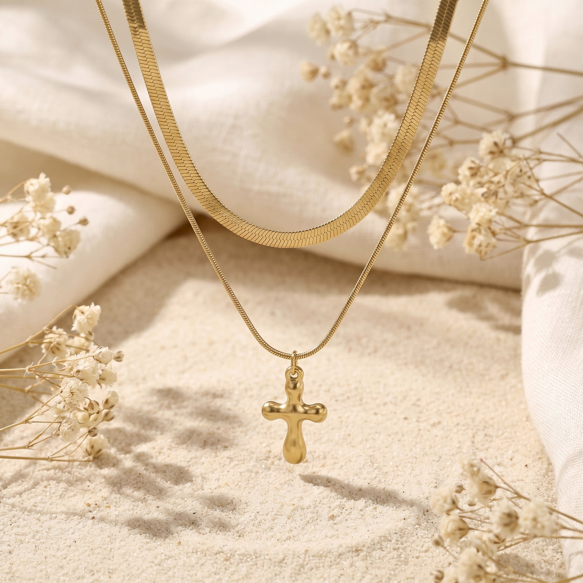 Gold-tone layered cross necklace displayed over sand, white fabric, and dried flowers.