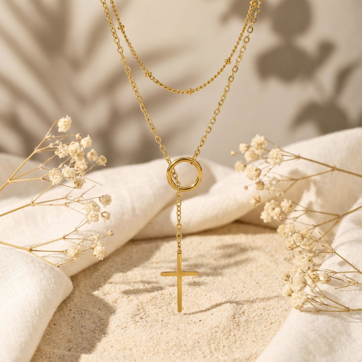 Gold-tone layered lariat necklace displayed vertically over sand and linen, with a ring station and cross pendant.