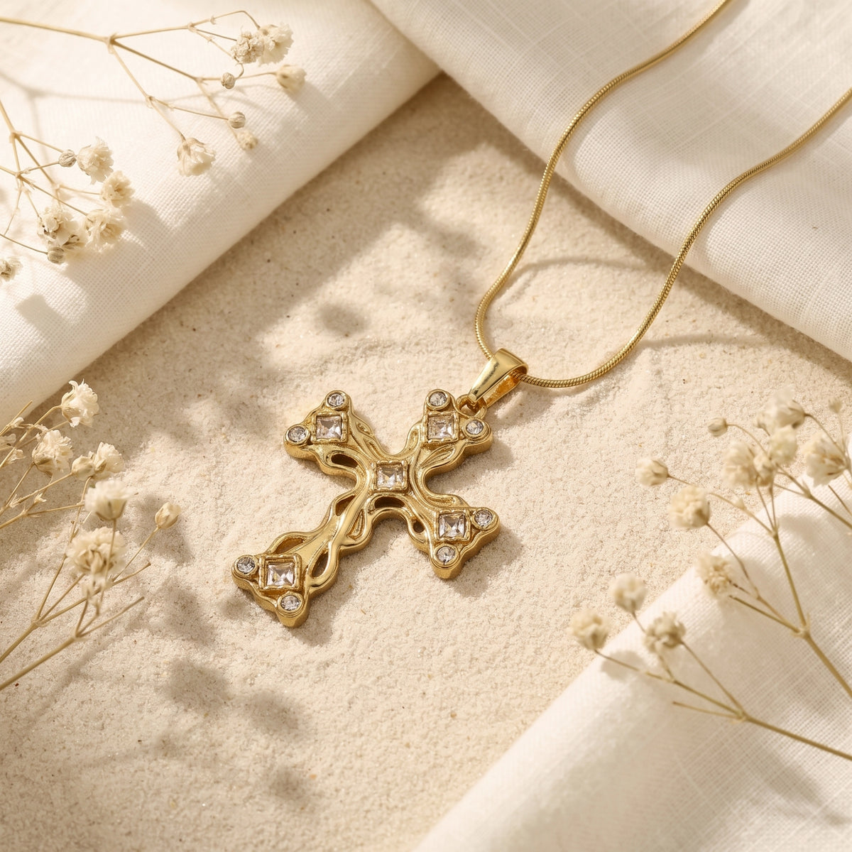 Gold-tone filigree cross necklace resting diagonally on sand beside white fabric and dried flowers.