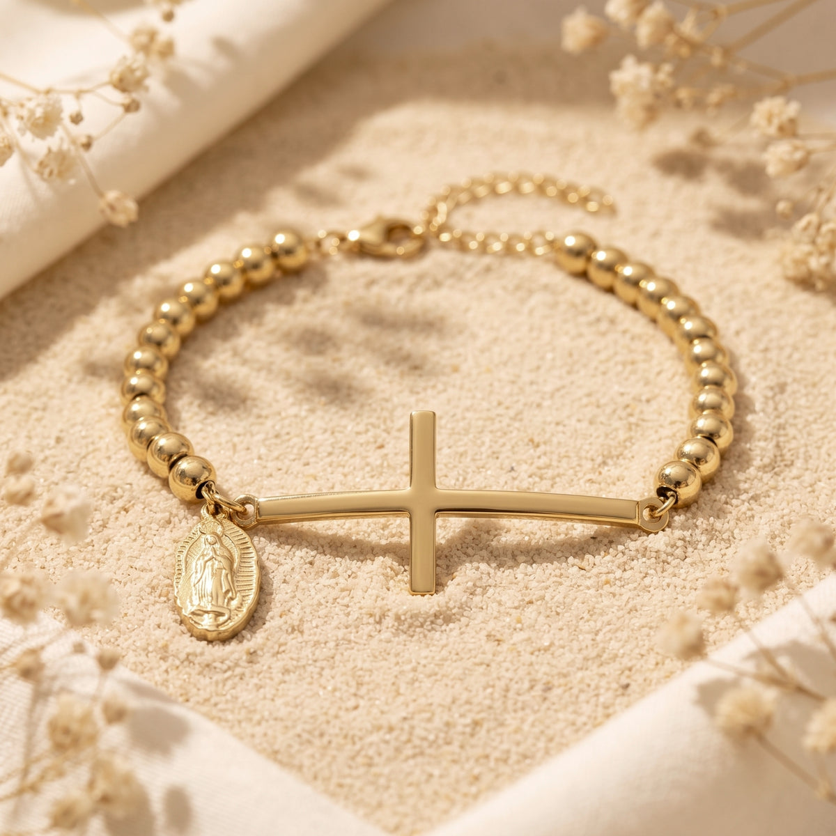 Detailed view of the gold-tone horizontal cross and Virgin Mary medal charm on a beaded bracelet resting on sand.