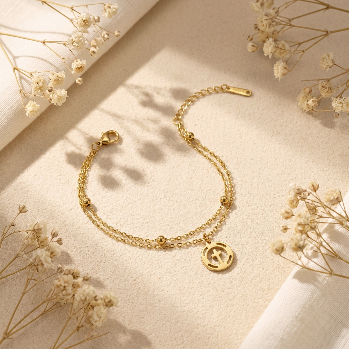 Gold-tone double-strand cross bracelet laid flat on sand, showing its clasp and extender chain.