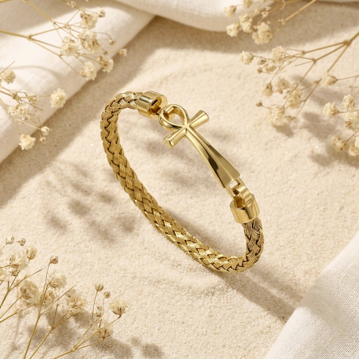 Angled view of the gold-tone braided Ankh bracelet on sand, showing the clasp and woven band.