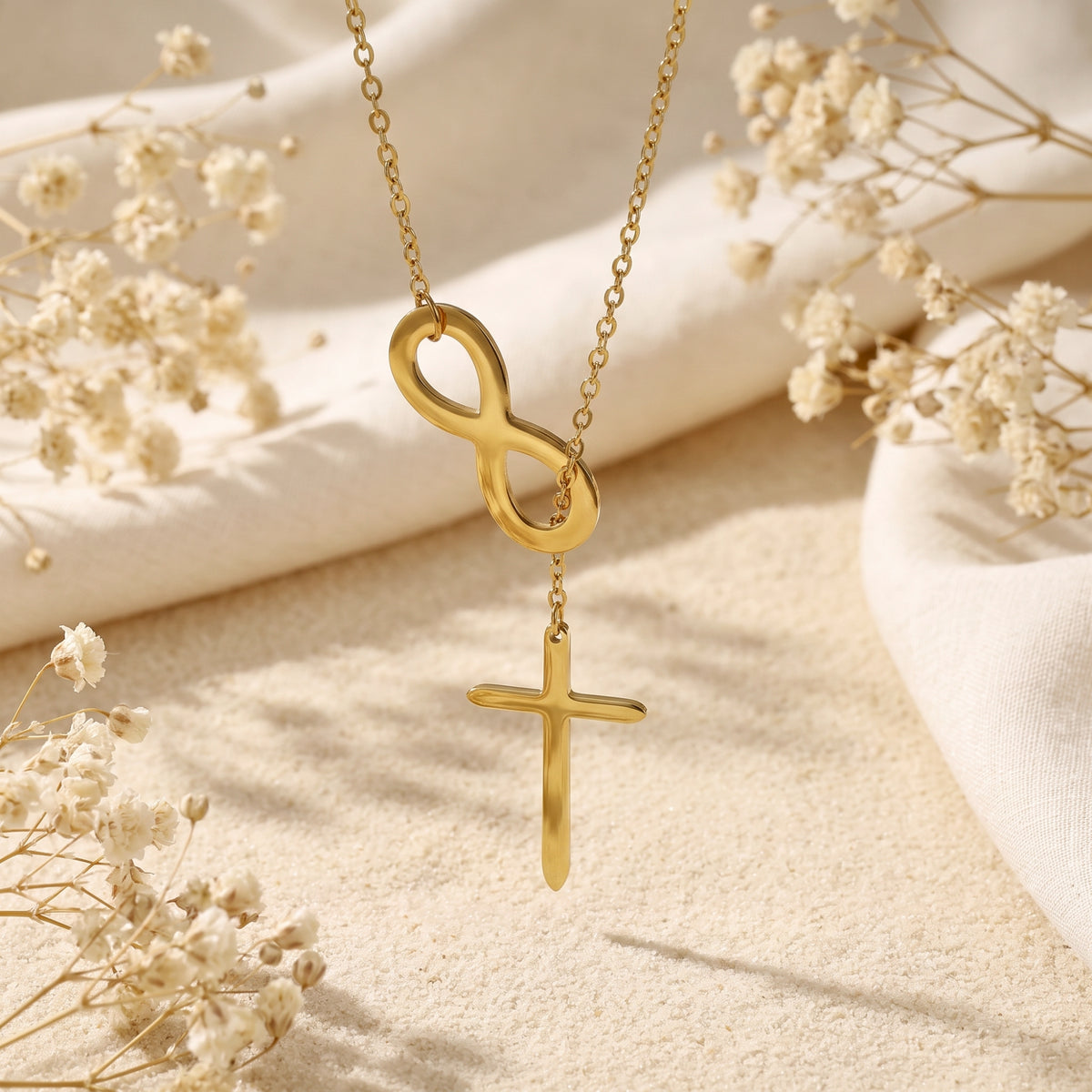 Gold-tone infinity and cross lariat necklace suspended over a textured beige surface with dried flowers