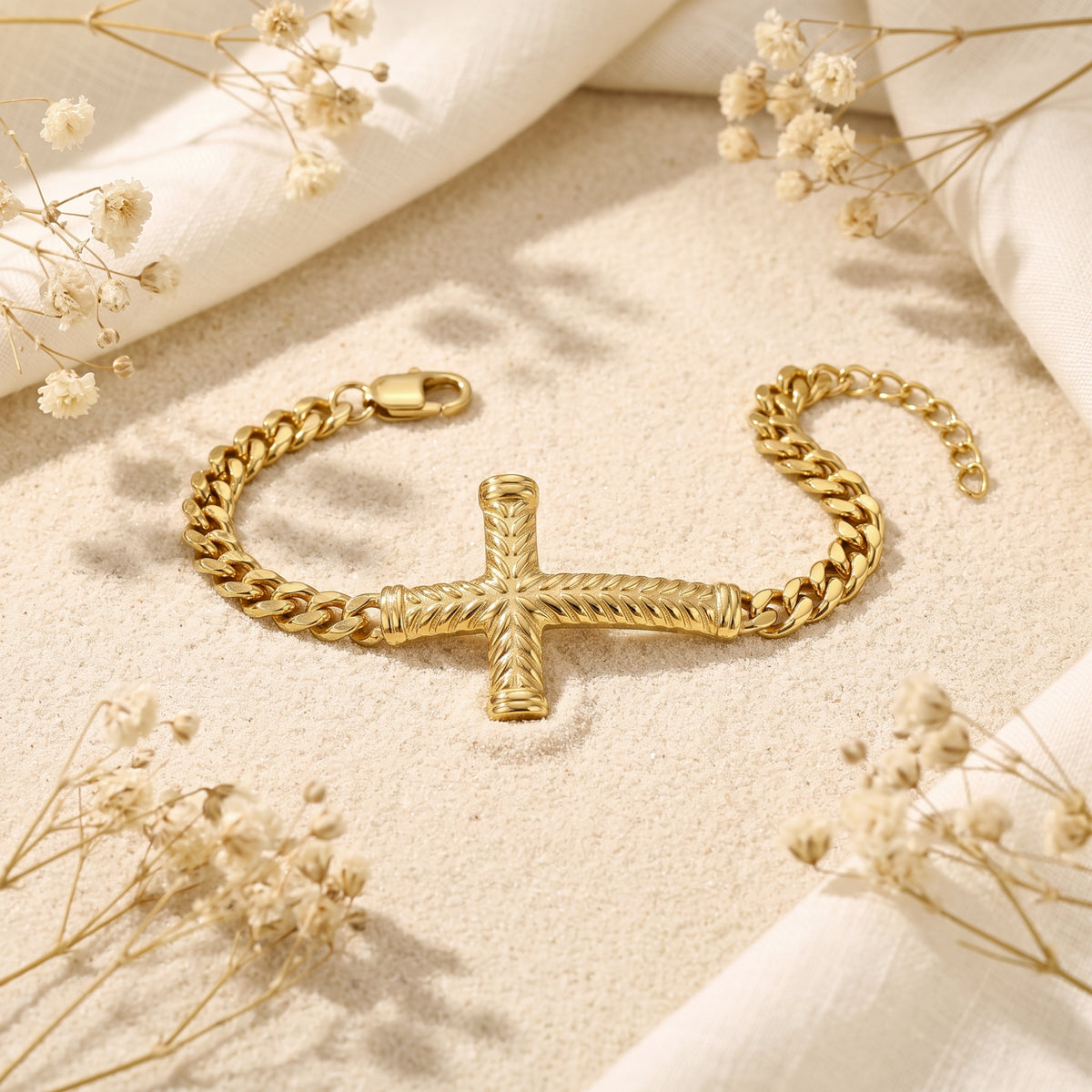 Unclasped gold-tone Cuban-link bracelet laid flat on a sandy surface, with a chevron-textured cross centered.