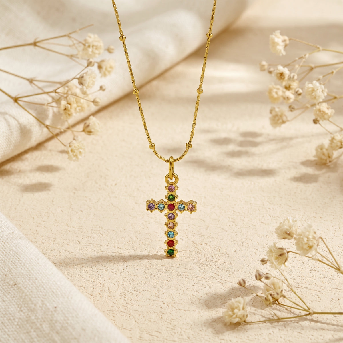 Gold-tone cross necklace displayed vertically on pale sand among dried white flowers.