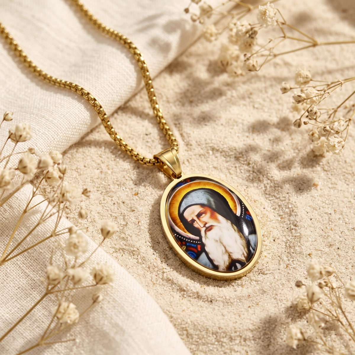 Gold-tone oval saint pendant lying flat on sand beside white fabric and dried flowers.