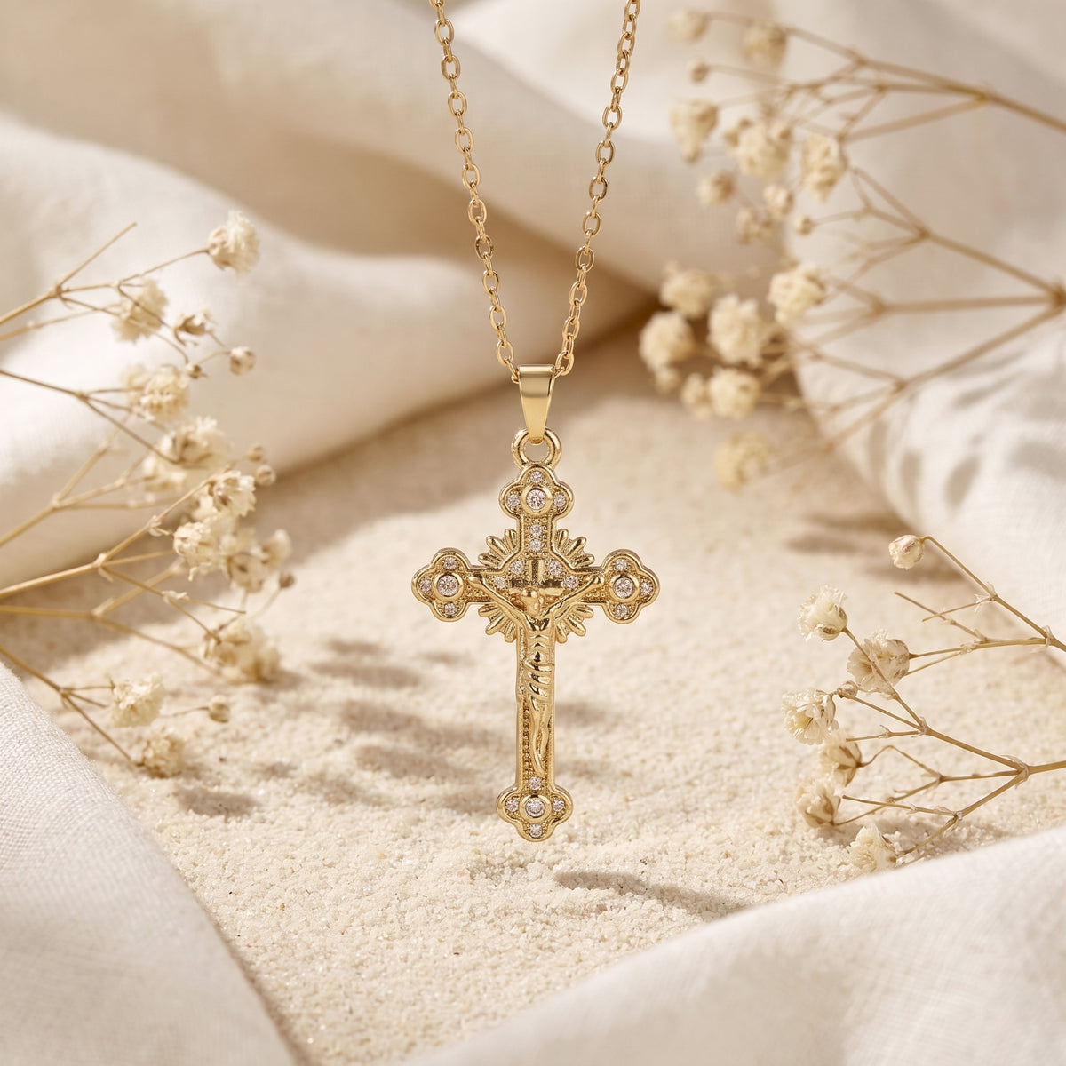 Front view of the gold-tone ornate cross pendant with clear accents hanging above sand and cream fabric.
