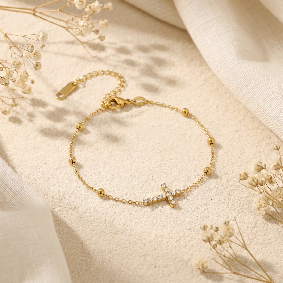 Gold-tone pave cross bracelet arranged in a circle on sand, with its clasp and extender chain visible.