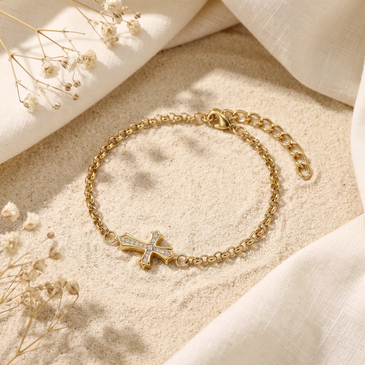 Angled flat lay of a gold-tone cross bracelet showing its clasp and adjustable extension chain on sand.