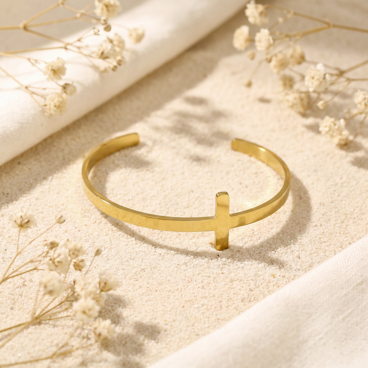 Gold-tone cross cuff bracelet lying flat on pale sand among dried flowers and cream fabric.