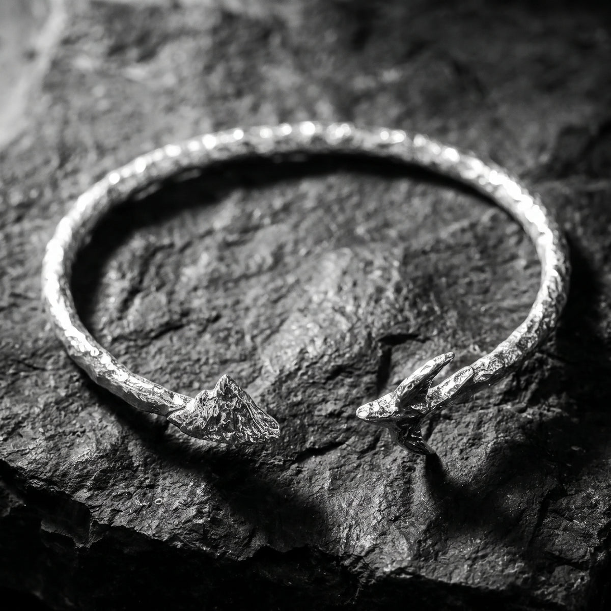 Overhead view of a textured silver open cuff bracelet resting on dark rock, with mountain and bird ends facing inward.
