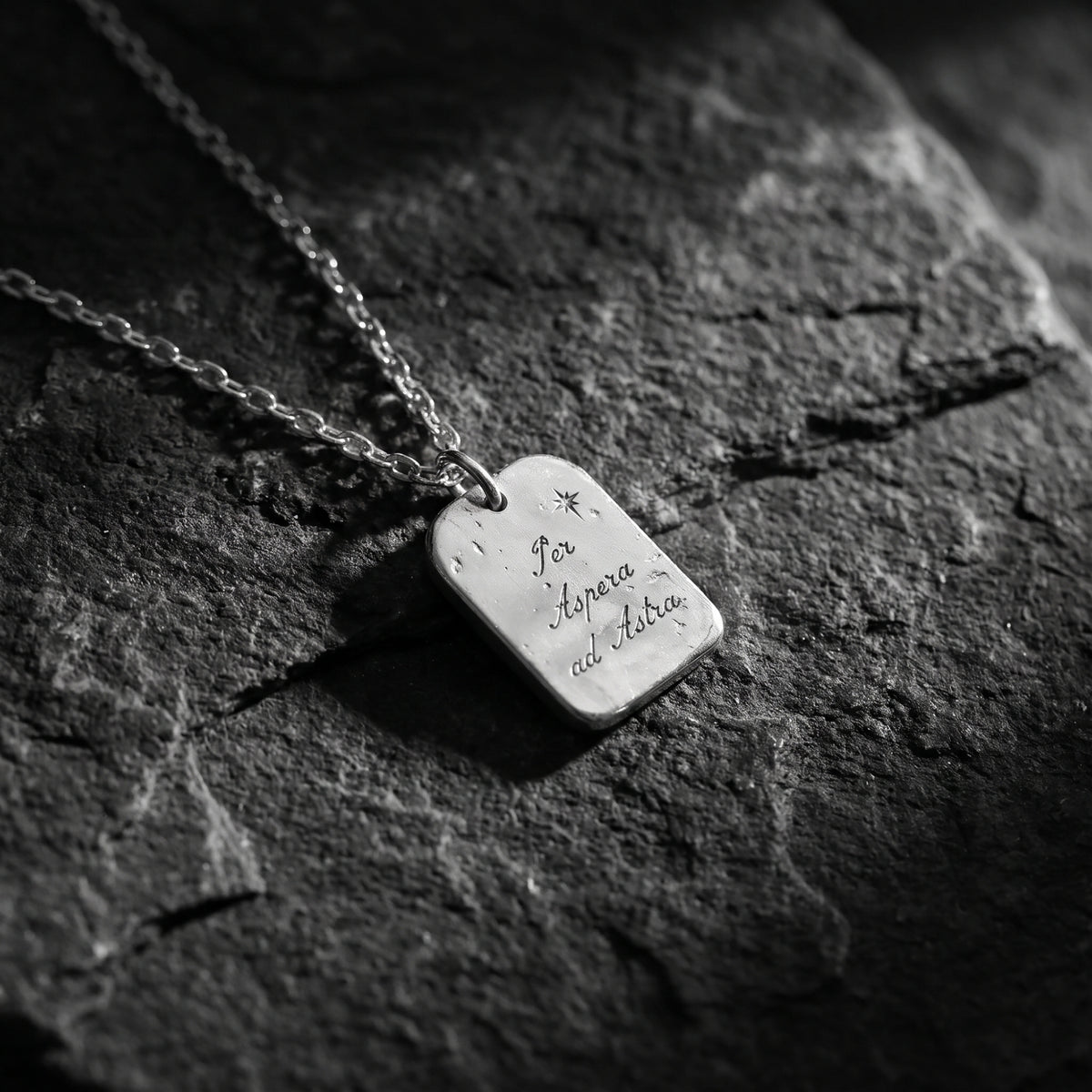 Angled close-up of the silver engraved tag pendant with star detail lying flat on dark stone.