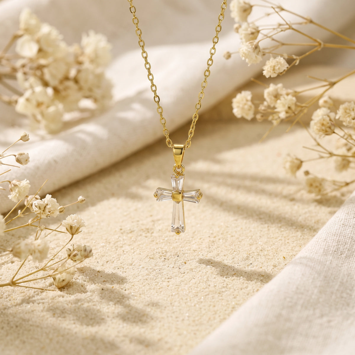 Gold cross necklace hanging vertically over sand, surrounded by white fabric and dried flowers.