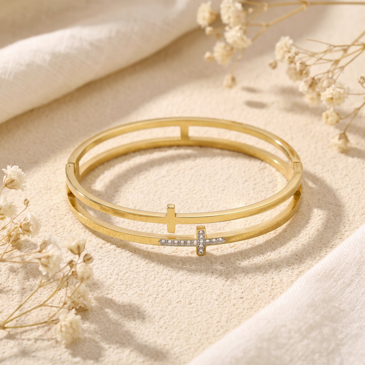 Gold-tone double-band cross bangle lying open on beige textured fabric with dried flowers.