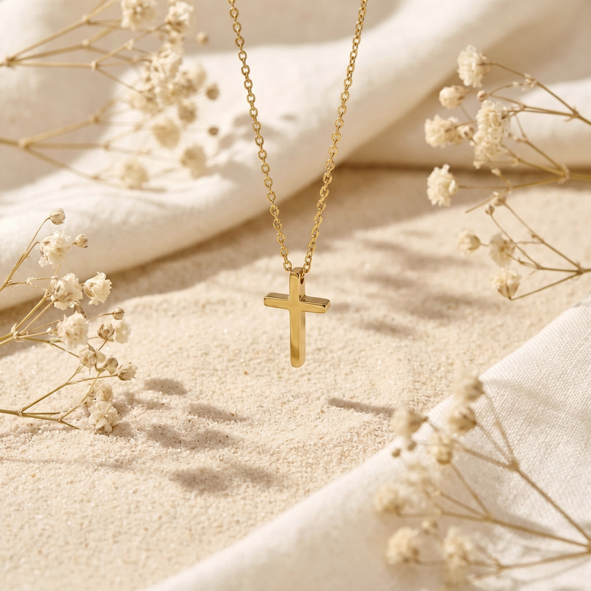 Gold-tone cross pendant necklace hanging vertically above sand, surrounded by dried white flowers and cream fabric.