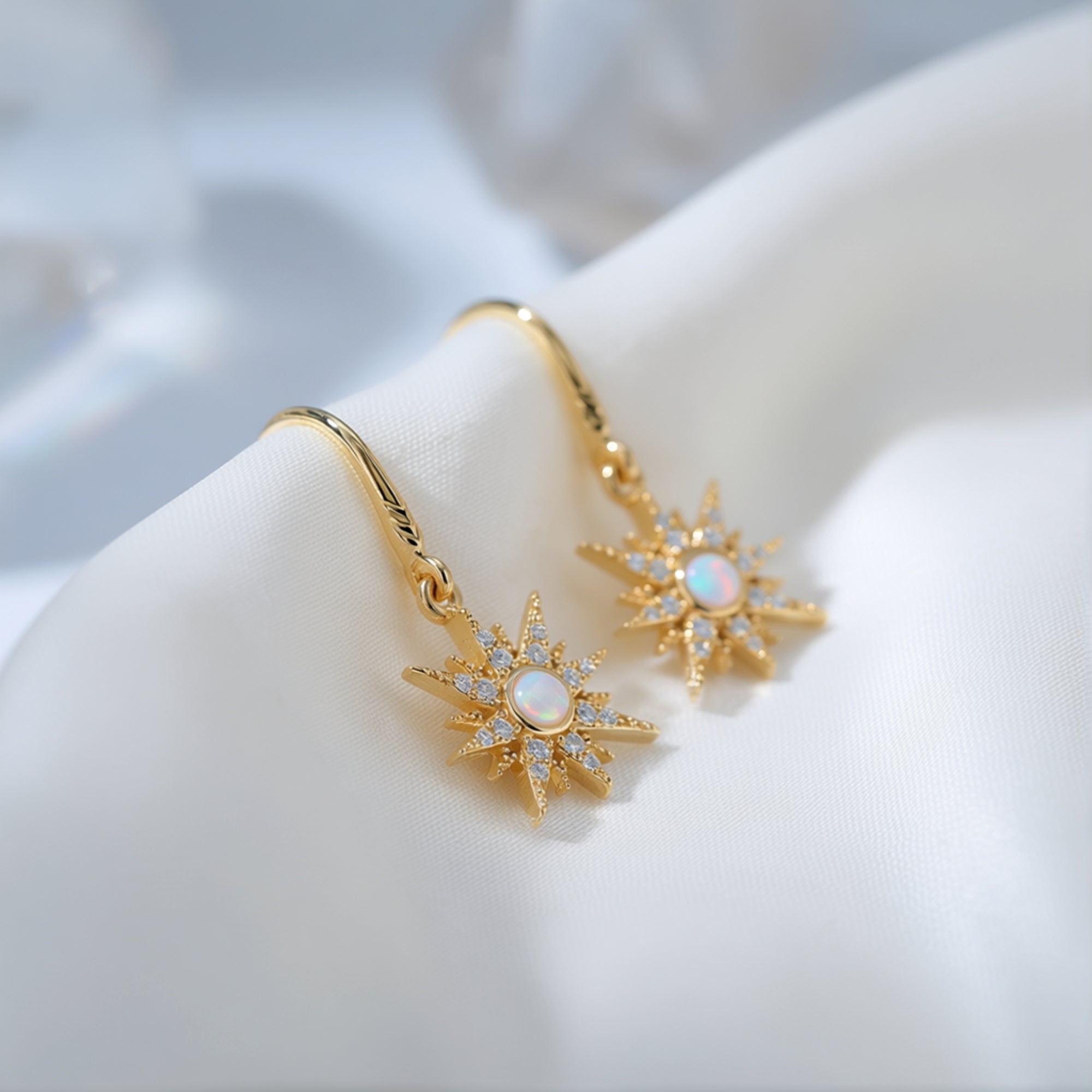 Close-up of gold-tone starburst drop earrings with round white opals and accent stones on white fabric.