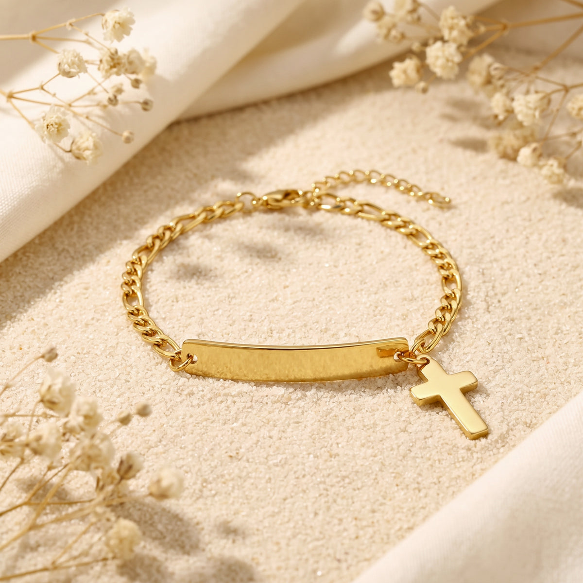 Close-up of the gold-tone Figaro bracelet showing its blank ID bar, cross charm, clasp, and extender chain on sand.