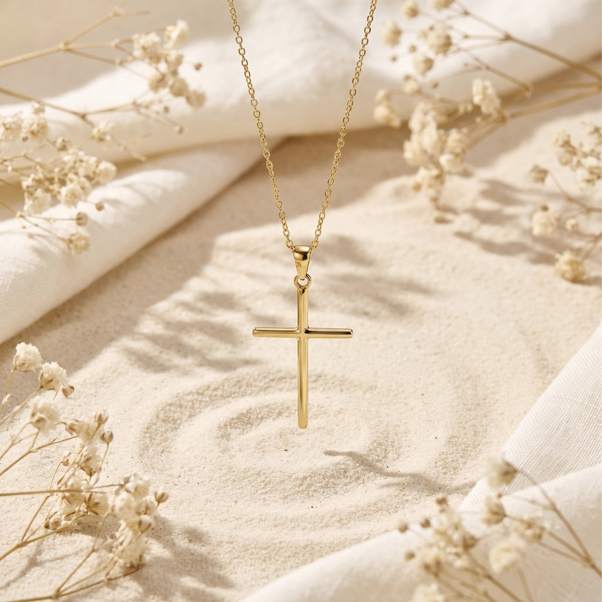 Gold-tone cross necklace suspended vertically over sand, framed by white fabric and dried flowers.