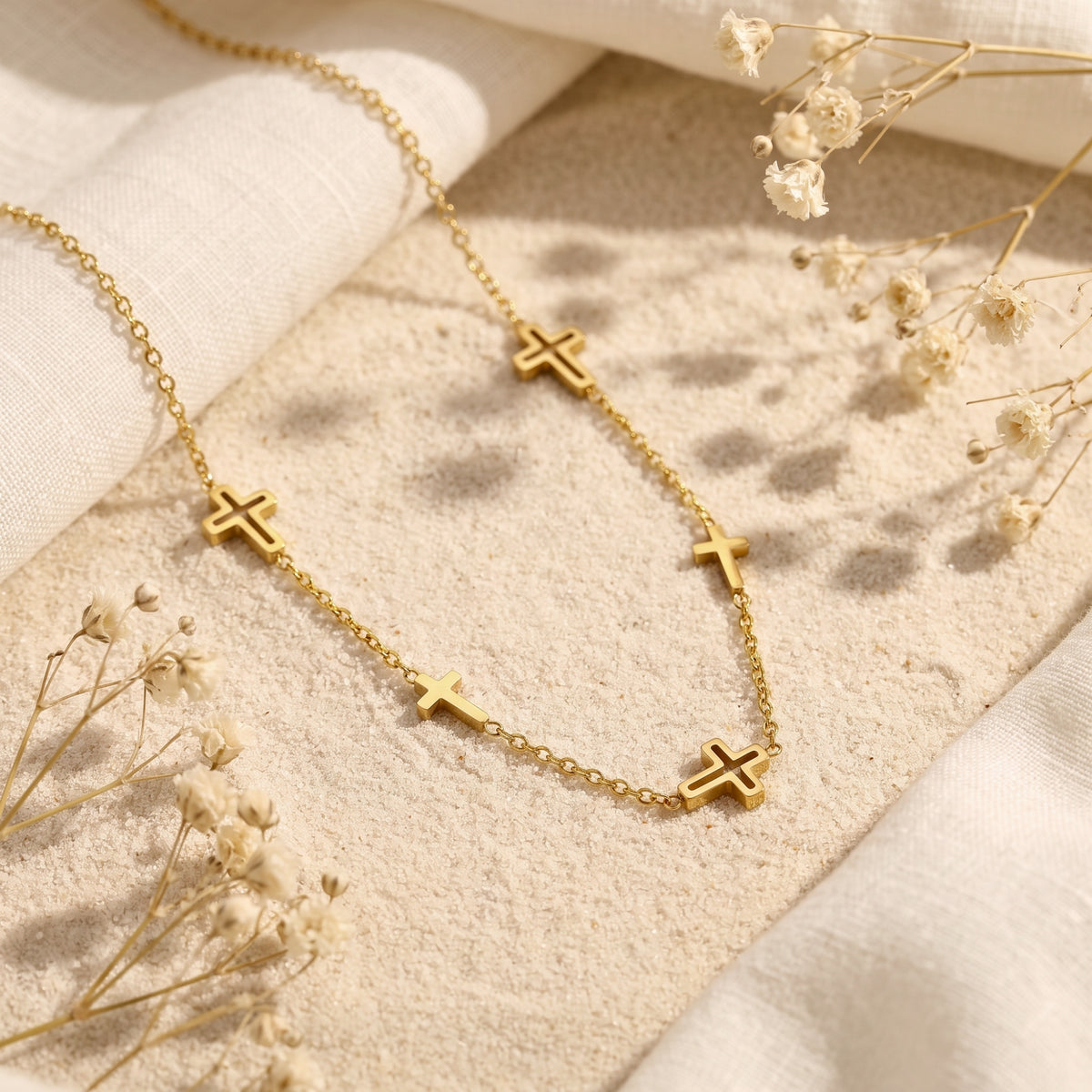 Gold-tone multi-cross station necklace laid diagonally on textured sand with dried flowers and white fabric.