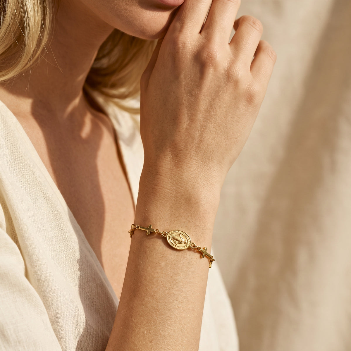 Close-up of a gold-tone cross station bracelet on a model's wrist as her hand rests near her face.