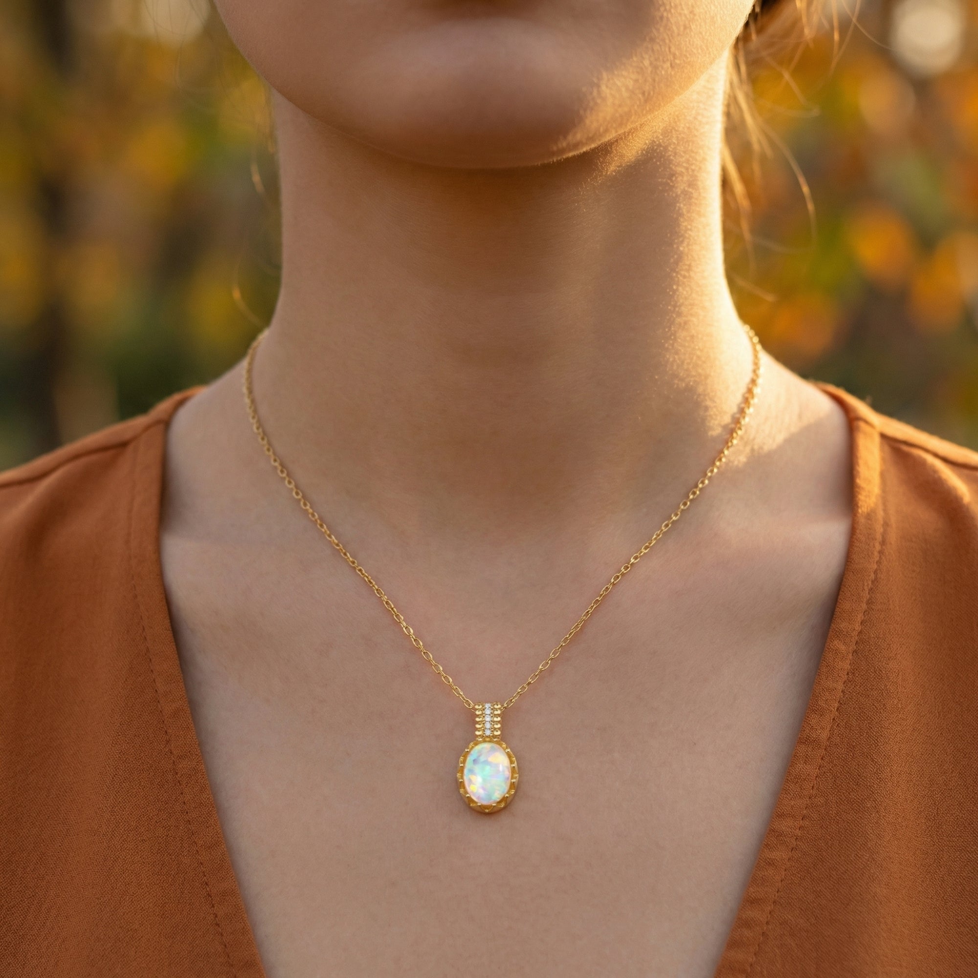 Frontal view of a model wearing the gold-tone Orianthi necklace with an oval white opal pendant and orange top.