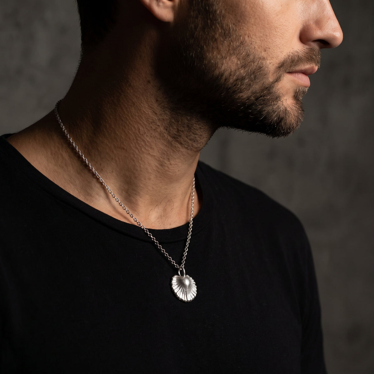 Side-profile man wearing the silver scallop shell pendant over a black crewneck shirt.
