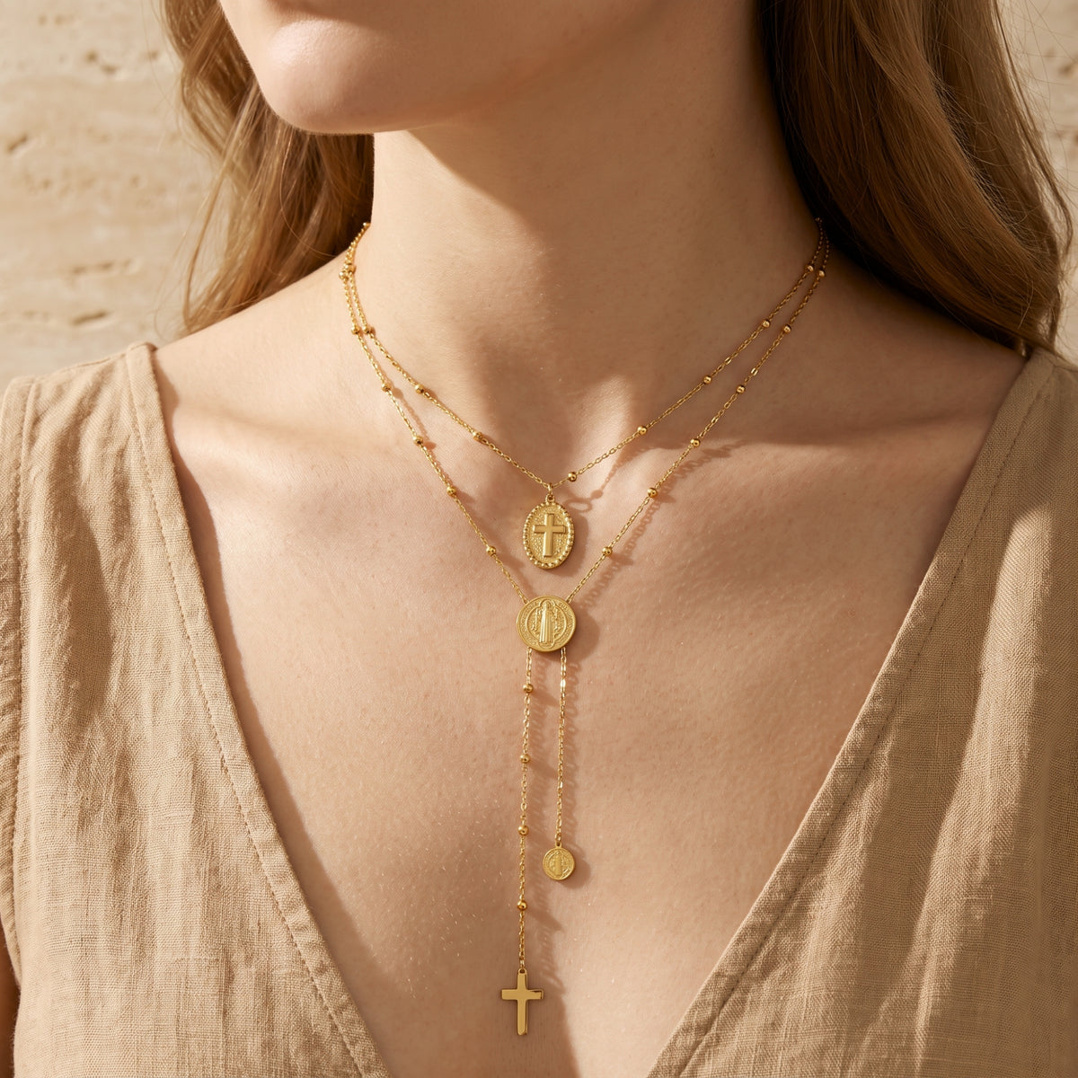 Front view of the gold-tone layered necklace with cross medallion and dangling cross worn with a beige V-neck garment