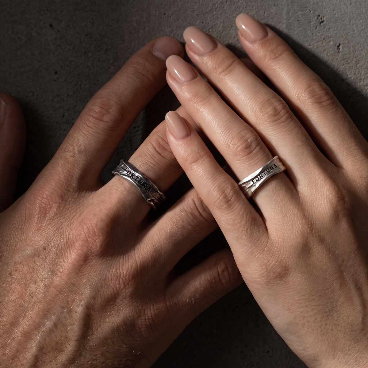 Top-down close-up of two overlapping hands wearing textured open silver rings stamped FORTUNA.