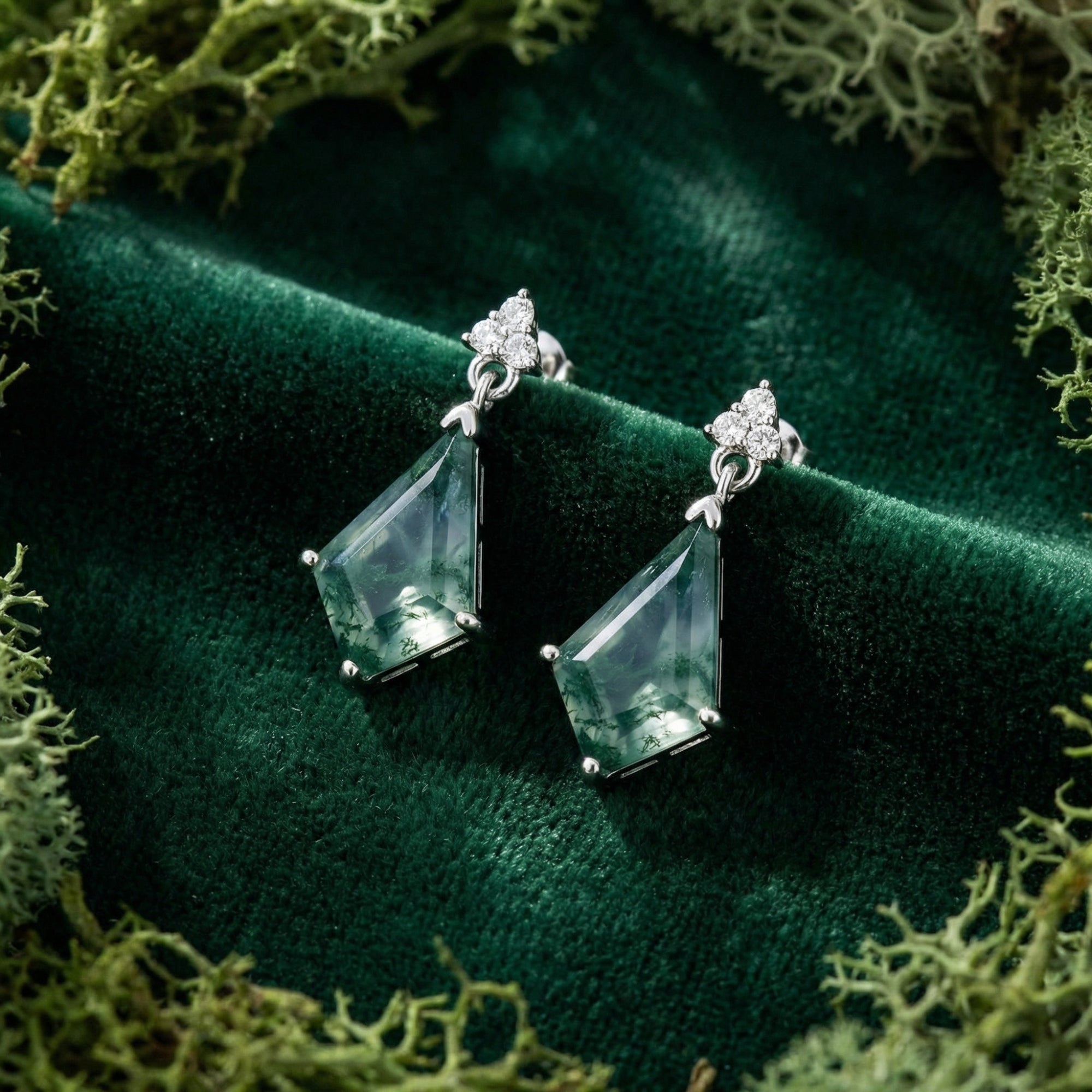 Pair of silver kite-cut moss agate drop earrings displayed on a textured green velvet surface.