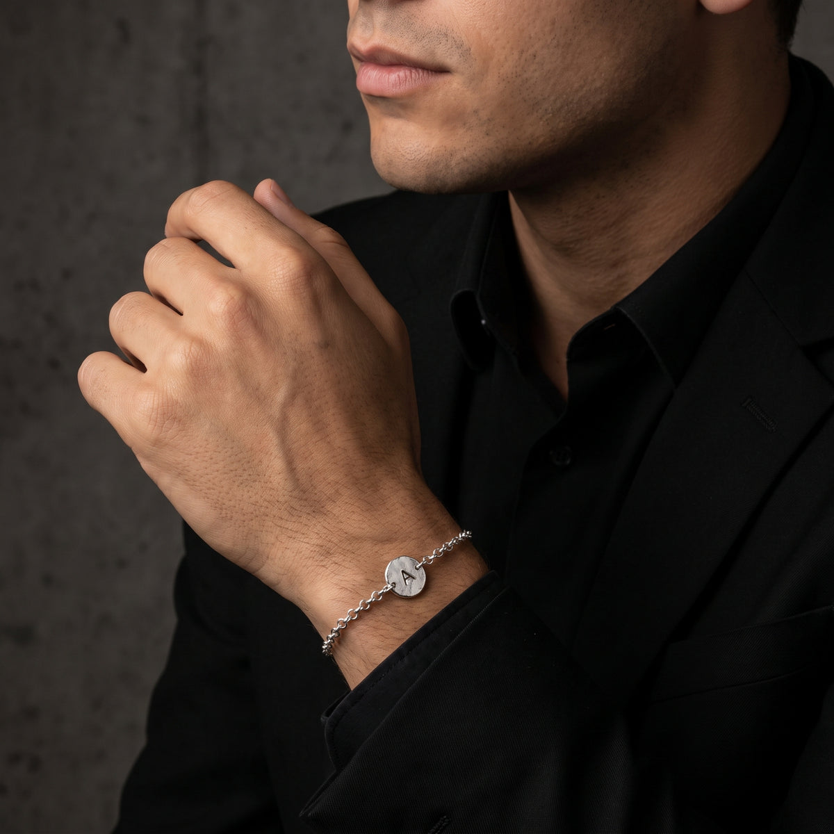 Close-up of a man wearing the silver initial A bracelet near his chin with a black suit and shirt.