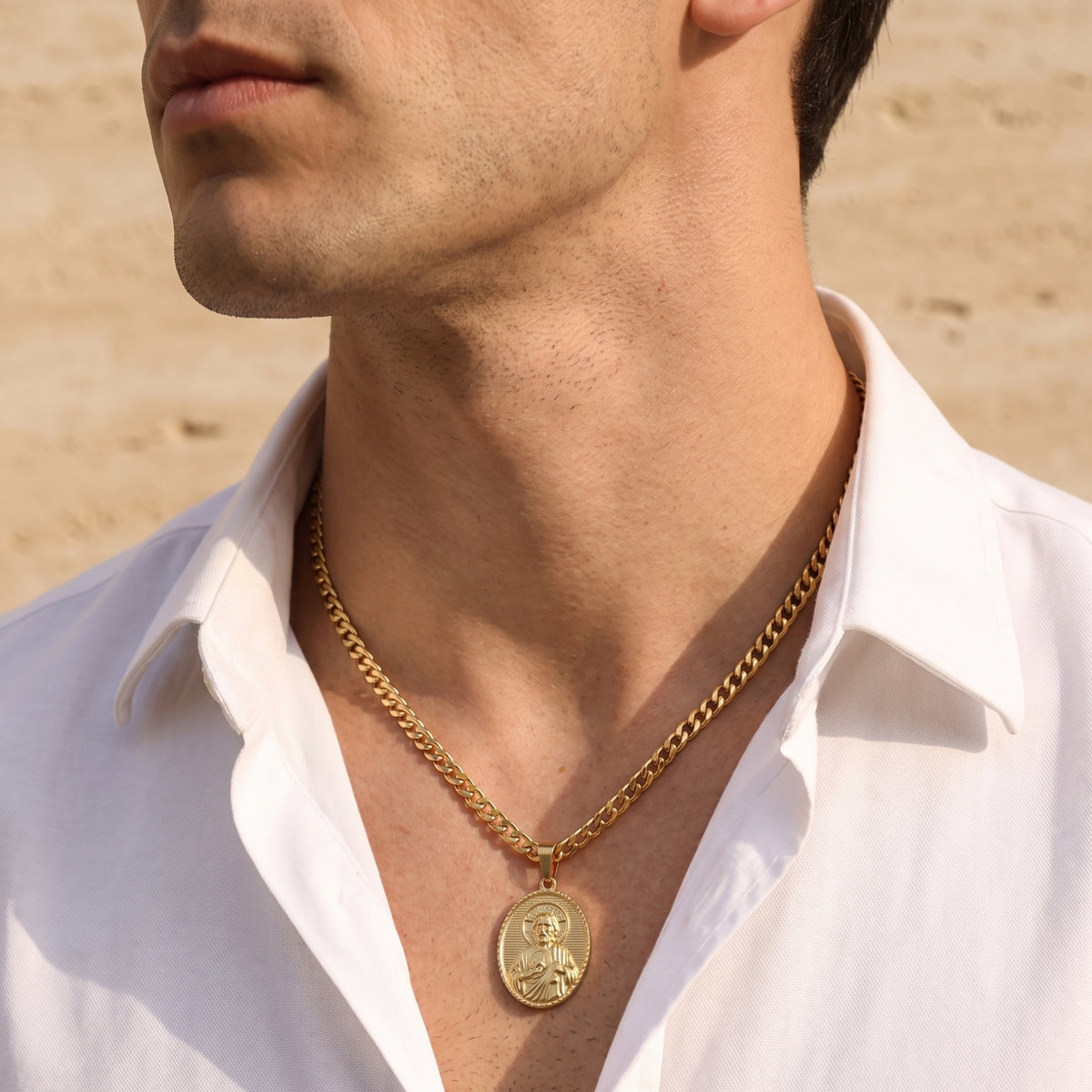 Close-up of a man wearing a gold-tone oval Saint Jude pendant necklace with a white shirt and sandy background.