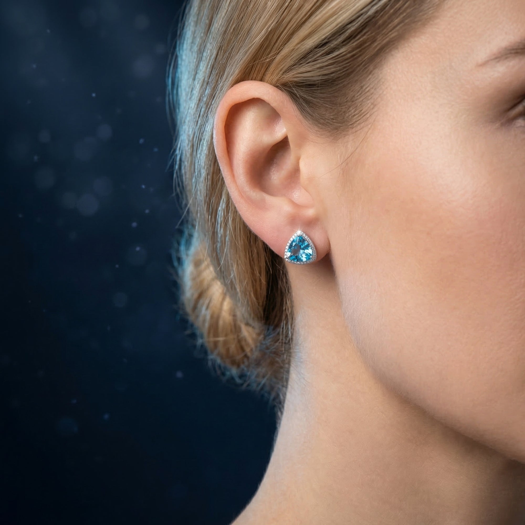 Side view of a woman wearing a silver triangle cut blue topaz earring with a halo of accent stones.
