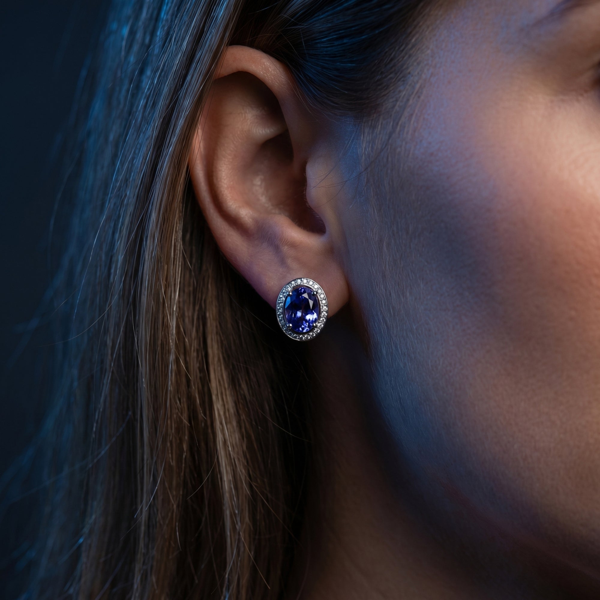 Model wearing an 18K white gold oval tanzanite stud earring with a diamond halo in soft lighting.