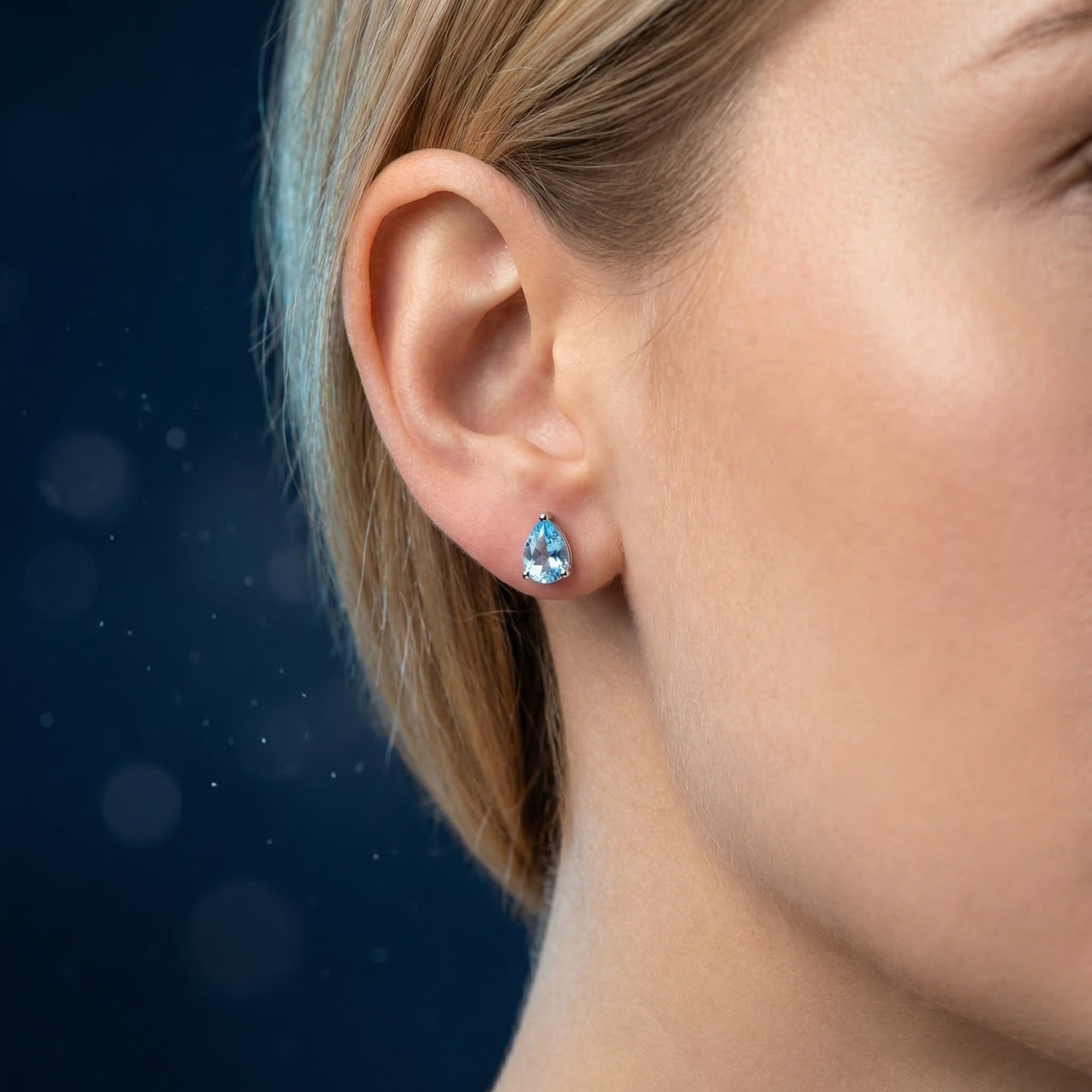 Close-up of a woman wearing a pear-cut natural blue topaz earring in a white gold setting against a dark background.