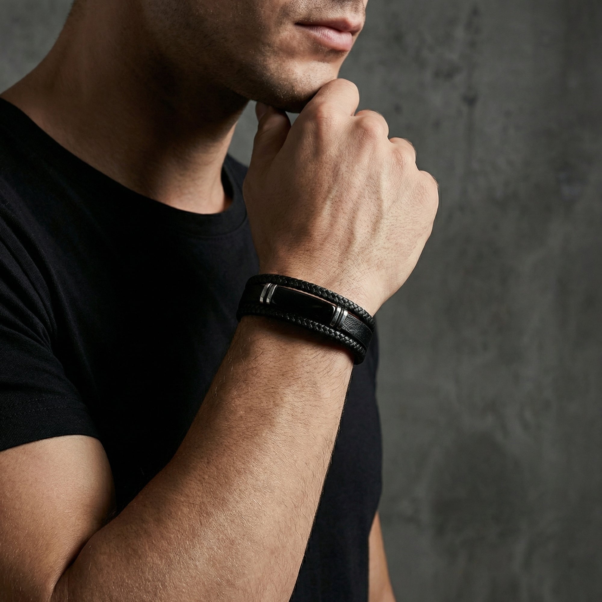 Model wearing the black braided leather Phantom Bracelet with a black stainless steel clasp against a grey wall.