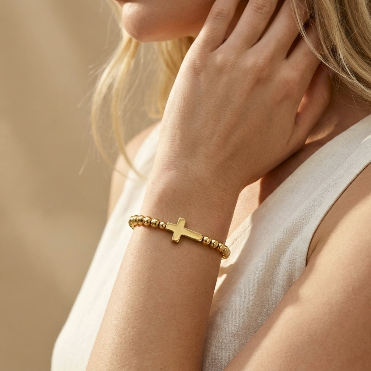 Model wearing a gold-tone beaded cross bracelet on her wrist with her hand touching her neck.