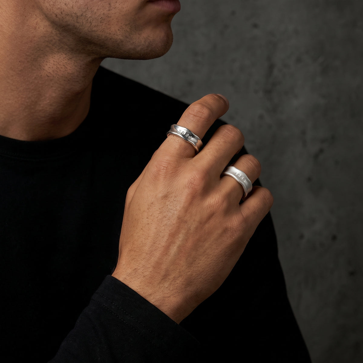 Man wearing two stamped silver open-band rings on separate fingers with his hand raised near his face